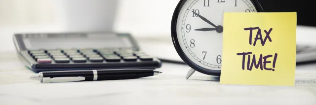 Sticky note with Tax Time! written on it on a desk with a clock, pen and calculator