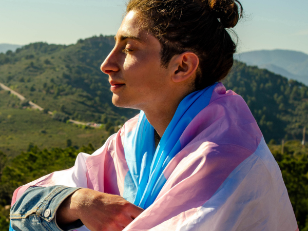 Transgender person wrapped in a Trans flag sitting on a mountain