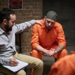 Caucasian man in orange prison uniform sitting with head down being comforted by middle aged Caucasian man holding clipboard during counseling session in prison